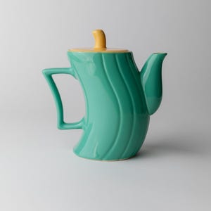 May include: A teal ceramic teapot with a yellow lid and handle. The teapot has a curved body with vertical wave-like ridges. The handle is a rectangular shape, and the spout is curved.