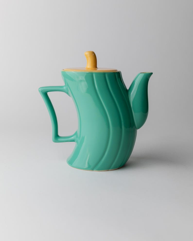 May include: A teal ceramic teapot with a yellow lid and handle. The teapot has a curved body with vertical wave-like ridges. The handle is a rectangular shape, and the spout is curved.