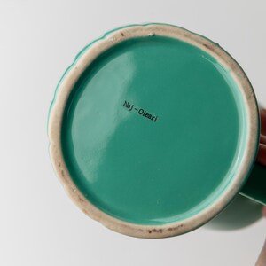 May include: Bottom view of a teal ceramic mug with a scalloped rim and a green handle. The interior is a solid teal color, and the text "Naj-Olear" is printed on the bottom.