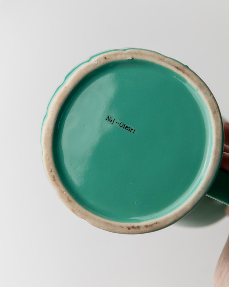 May include: Bottom view of a teal ceramic mug with a scalloped rim and a green handle. The interior is a solid teal color, and the text "Naj-Olear" is printed on the bottom.