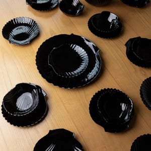Könnte beinhalten: Eine Sammlung von glänzend schwarzem Geschirr in Form von Jakobsmuscheln, bestehend aus Tellern und Schalen in verschiedenen Größen mit gerippten Rändern, die verstreut auf einer hellen Holzoberfläche angeordnet sind.