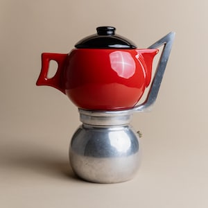 Könnte beinhalten: Eine rote und silberne Espressomaschine für den Herd mit einem schwarzen Deckel. Die Maschine hat einen runden, roten Topf auf einem silbernen Sockel.