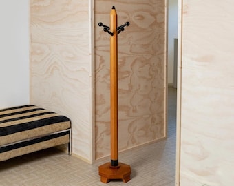 Può includere: Un attaccapanni in legno a forma di matita gigante. L'attaccapanni a forma di matita ha una punta e una gomma nere, con pioli neri per appendere i cappotti. La base è esagonale. Si trova in una stanza con pannelli di legno chiaro.