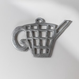 Könnte beinhalten: Ein silberfarbener Metall-Topfuntersetzer in Form einer Gießkanne. Der Untersetzer hat einen Griff, einen Ausguss und ein gitterartiges Muster in der Mitte. Er dient zum Schutz von Oberflächen vor heißen Gegenständen.