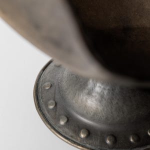 May include: Close-up of a bronze-colored metal bowl with a rounded base and a flared rim. The base features a series of evenly spaced rivets. The bowl has a weathered, antique appearance.