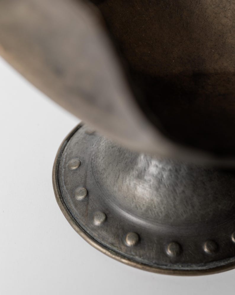 May include: Close-up of a bronze-colored metal bowl with a rounded base and a flared rim. The base features a series of evenly spaced rivets. The bowl has a weathered, antique appearance.