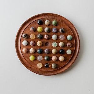 May include: A circular wooden solitaire board game featuring a rich brown grain and a series of indentations. The board is populated with various polished, multi-colored stone spheres in shades of green, black, white, yellow, and brown.