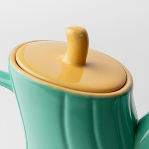 May include: Close-up of a teal ceramic teapot with a yellow lid and handle. The teapot has a curved design and a glossy finish. The lid has a small, rounded handle. The background is plain white.
