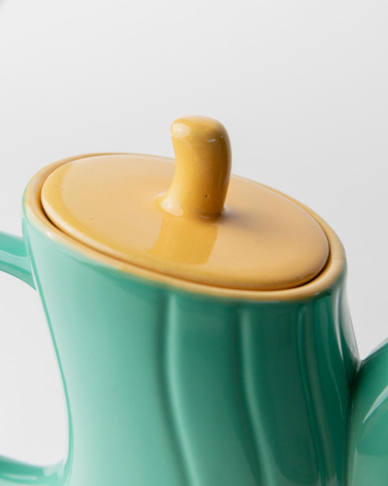 May include: Close-up of a teal ceramic teapot with a yellow lid and handle. The teapot has a curved design and a glossy finish. The lid has a small, rounded handle. The background is plain white.