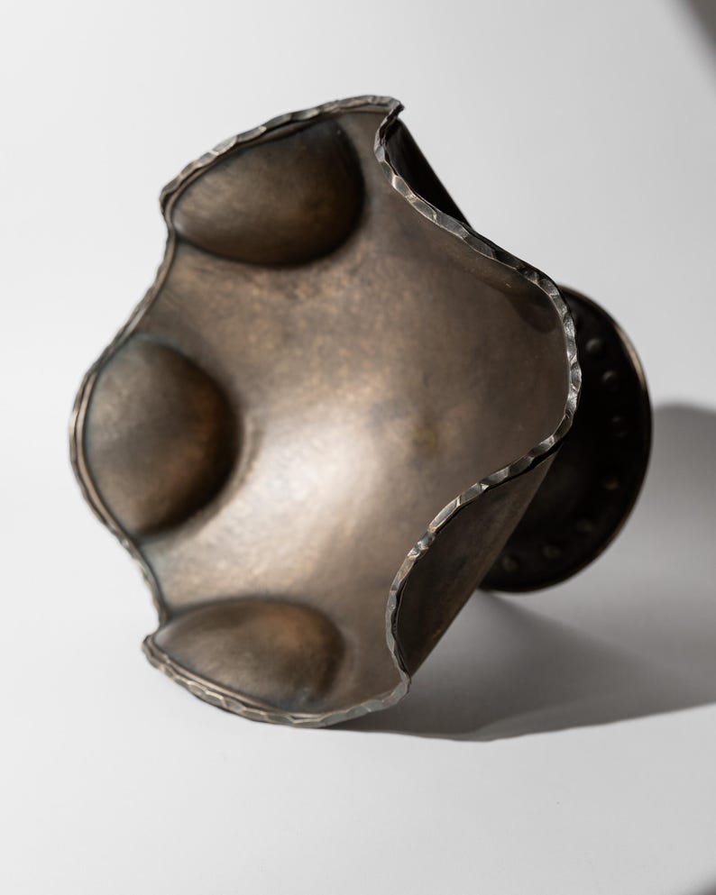 May include: A handcrafted, metallic bronze-colored bracelet with a unique, undulating design. The bracelet features a textured, hammered edge and three raised, rounded elements. The overall shape is organic and sculptural, suitable for artistic jewelry.