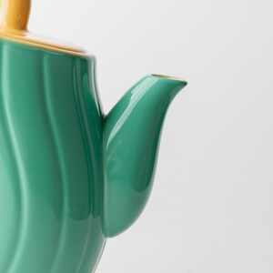 May include: Close-up of a ceramic teapot with a green body and a yellow lid and handle. The teapot has a curved spout and a ridged design on the body. The teapot is on a white background.