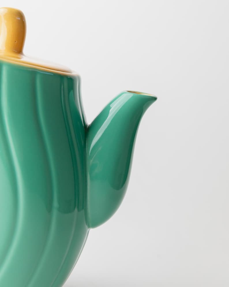 May include: Close-up of a ceramic teapot with a green body and a yellow lid and handle. The teapot has a curved spout and a ridged design on the body. The teapot is on a white background.