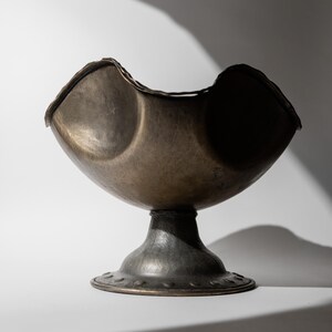 May include: A bronze-colored, hammered metal bowl with a pedestal base. The bowl has a unique, curved shape with two rounded depressions. The base is round with a decorative edge. The piece is likely a decorative item or a serving bowl.