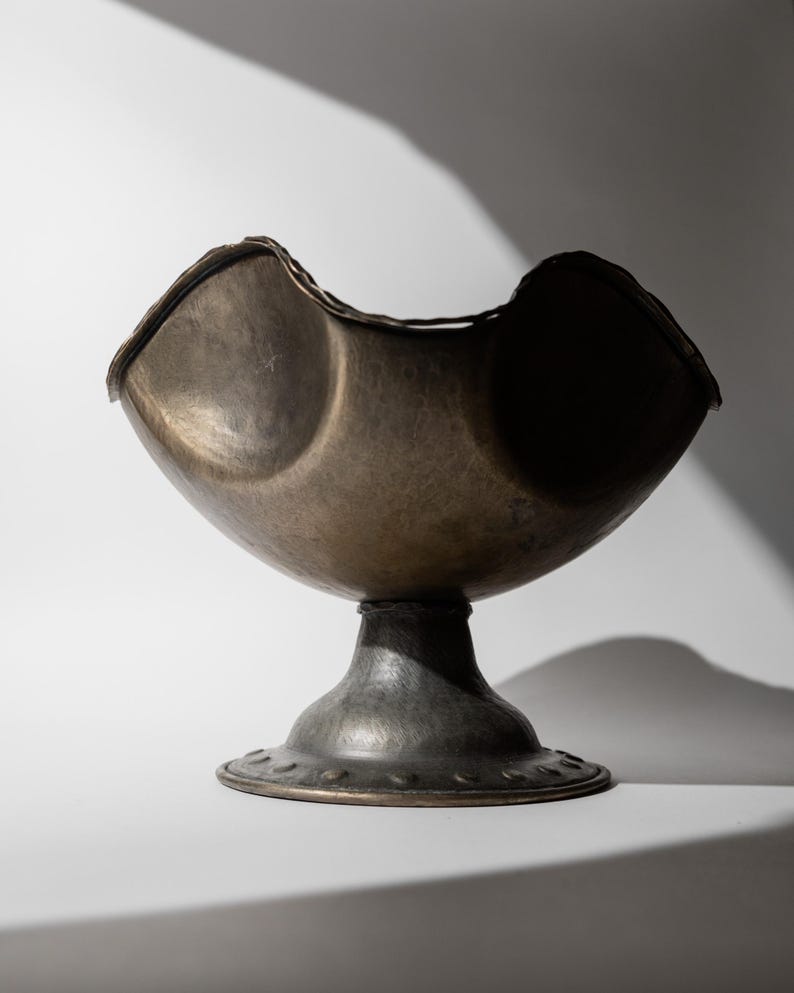 May include: A bronze-colored, hammered metal bowl with a pedestal base. The bowl has a unique, curved shape with two rounded depressions. The base is round with a decorative edge. The piece is likely a decorative item or a serving bowl.