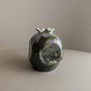 Könnte beinhalten: Eine Keramikvase in Form eines Granatapfels mit einer grün-blauen Glasur. Die Vase hat einen weißen Rand und eine strukturierte Oberfläche.