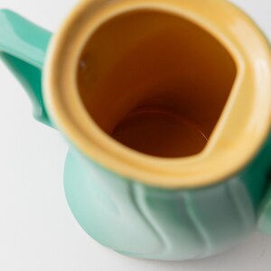 May include: A close-up of a ceramic pitcher with a light green exterior and a yellow-orange interior. The pitcher has a handle and a spout. The interior is a contrasting color to the exterior. The pitcher is on a white background.