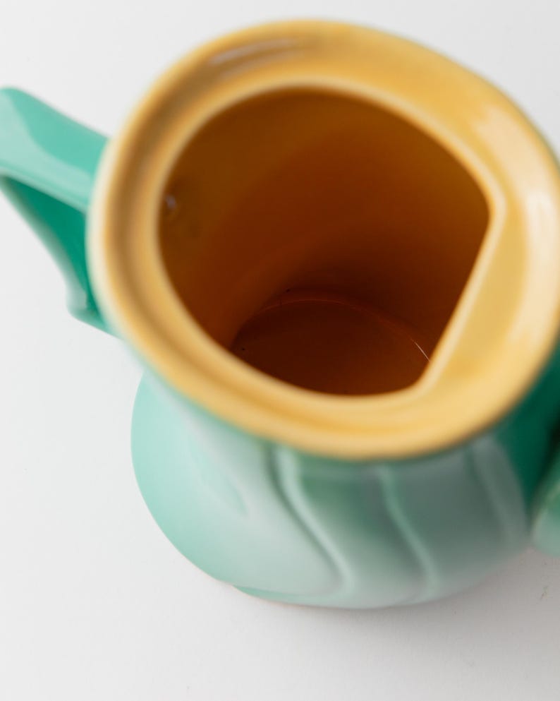 May include: A close-up of a ceramic pitcher with a light green exterior and a yellow-orange interior. The pitcher has a handle and a spout. The interior is a contrasting color to the exterior. The pitcher is on a white background.