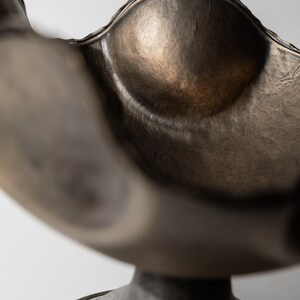 May include: A bronze-colored, decorative bowl with a scalloped rim and a rounded, textured interior. The bowl sits atop a pedestal base, creating a sculptural, artistic piece. The metal has a hammered appearance, adding to its unique aesthetic.