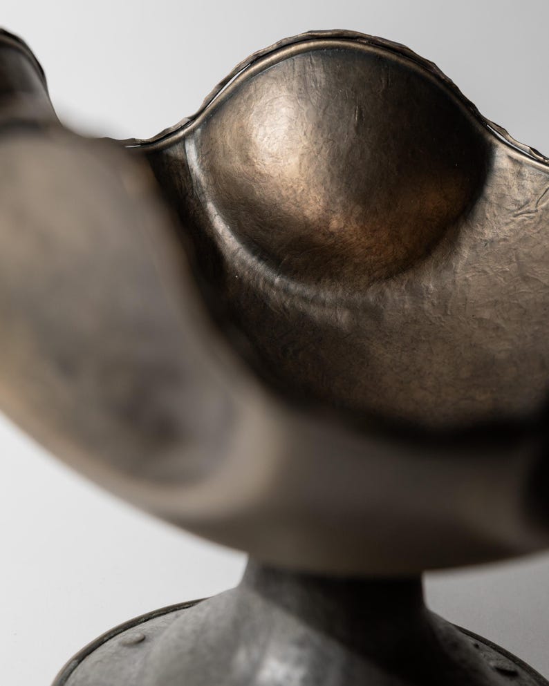 May include: A bronze-colored, decorative bowl with a scalloped rim and a rounded, textured interior. The bowl sits atop a pedestal base, creating a sculptural, artistic piece. The metal has a hammered appearance, adding to its unique aesthetic.