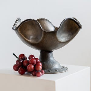 May include: A decorative metal bowl with a scalloped rim, resting on a pedestal base. The bowl has a dark, aged finish. A cluster of red grapes sits beside the bowl.