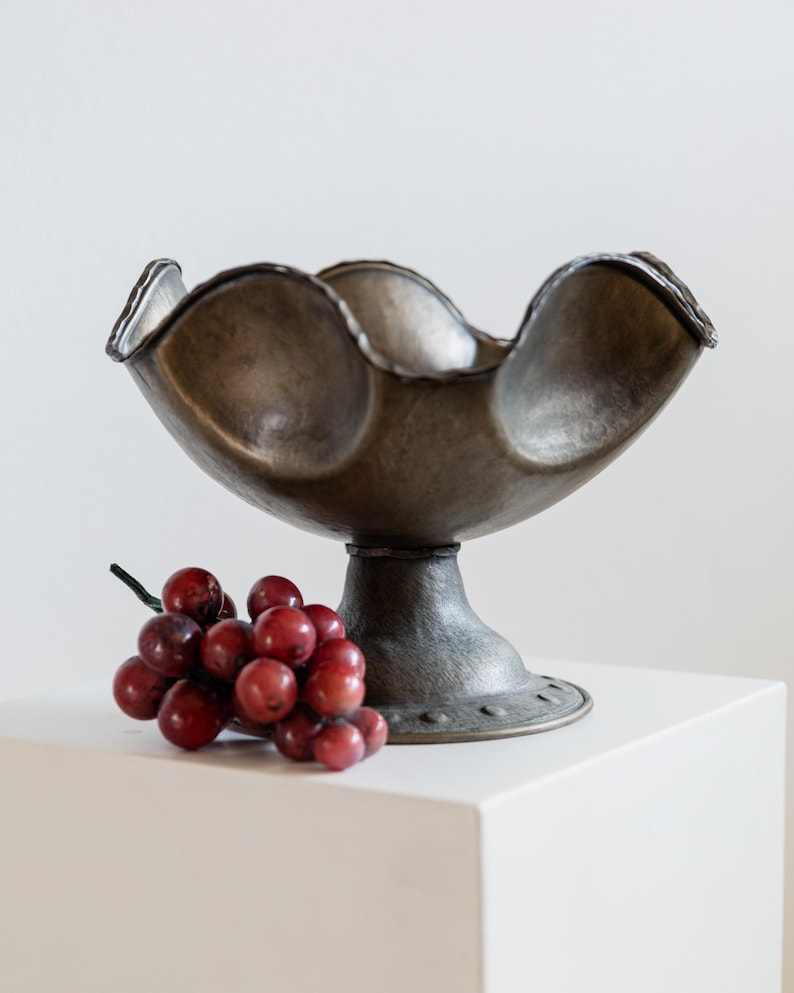 May include: A decorative metal bowl with a scalloped rim, resting on a pedestal base. The bowl has a dark, aged finish. A cluster of red grapes sits beside the bowl.