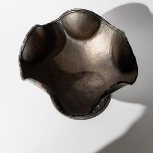 May include: A handcrafted, antique-style metal bowl with a unique, undulating rim. The bowl has a dark, metallic finish and a textured surface. The bowl is round with a pedestal base. The bowl is likely used for decorative purposes.