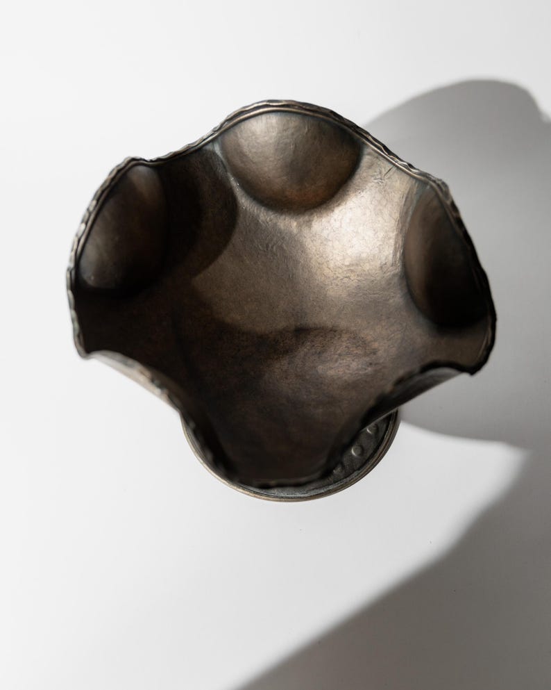 May include: A handcrafted, antique-style metal bowl with a unique, undulating rim. The bowl has a dark, metallic finish and a textured surface. The bowl is round with a pedestal base. The bowl is likely used for decorative purposes.