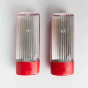 May include: Two clear, ribbed plastic containers with red bases. The containers are cylindrical and have a vertical, ridged design. The red bases have a rectangular indentation. The containers are likely for storing items.