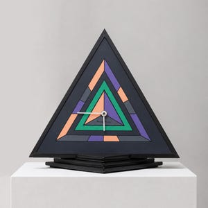 Peut inclure: Une horloge triangulaire avec un cadre noir et un cadran géométrique composé de triangles concentriques dans des tons de violet, vert, orange et gris foncé. L'horloge possède deux aiguilles argentées centrées sur le cadran, fixée sur un socle noir.