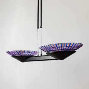 May include: A contemporary ceiling light with two striped glass shades. The shades have a blue, red, and white pattern. The light fitting has a black metal frame and is suspended from the ceiling.