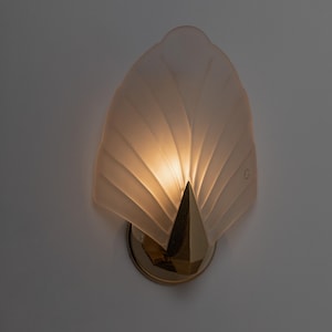 May include: A wall-mounted light fixture with a fan-shaped, frosted glass shade. The fixture has a gold-colored, geometric base and a pointed, gold-colored accent. The light is illuminated, casting a warm glow.