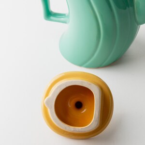 May include: A close-up of a teal ceramic teapot with a curved handle and a yellow lid. The lid is round with a white rim and a small hole in the center. The teapot has a smooth, glossy finish.