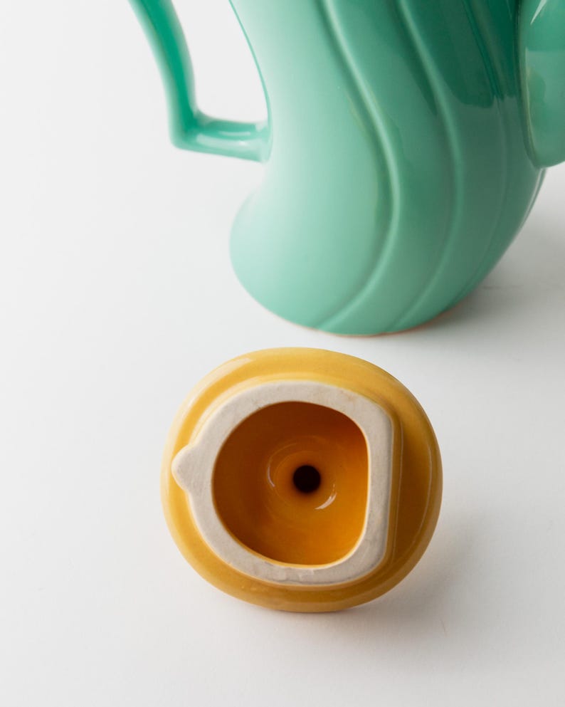May include: A close-up of a teal ceramic teapot with a curved handle and a yellow lid. The lid is round with a white rim and a small hole in the center. The teapot has a smooth, glossy finish.