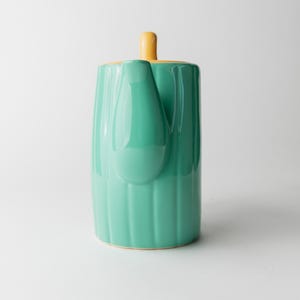 May include: A ceramic teapot in a vertical orientation, featuring a vibrant teal body with vertical ridged detailing. The lid and handle are a contrasting yellow. The teapot has a spout and a handle, and sits on a white surface.