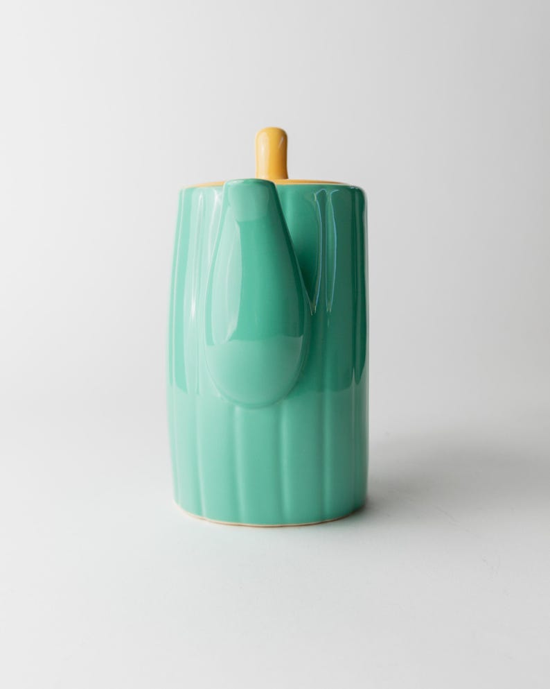 May include: A ceramic teapot in a vertical orientation, featuring a vibrant teal body with vertical ridged detailing. The lid and handle are a contrasting yellow. The teapot has a spout and a handle, and sits on a white surface.