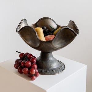 May include: A bronze-colored pedestal fruit bowl with a scalloped rim, filled with various fruits. A bunch of red grapes rests on the white surface beside the bowl. The bowl has a textured finish and a round base.