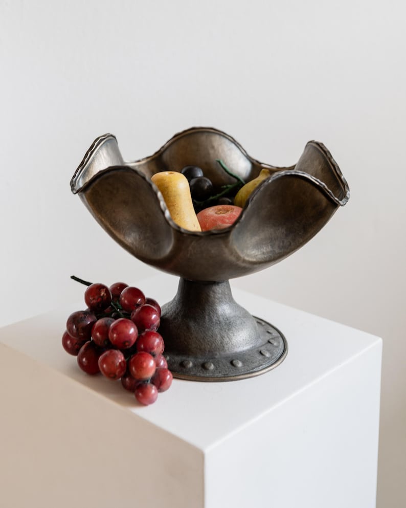 May include: A bronze-colored pedestal fruit bowl with a scalloped rim, filled with various fruits. A bunch of red grapes rests on the white surface beside the bowl. The bowl has a textured finish and a round base.