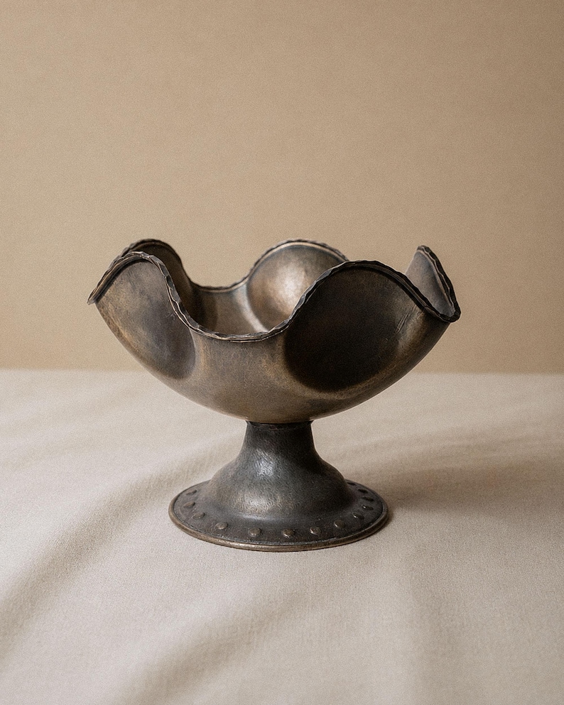 May include: A decorative metal bowl with a scalloped rim and pedestal base. The bowl has a dark, aged finish with a textured surface. The base features a series of small, evenly spaced indentations around the bottom edge. The bowl is empty and sits on a neutral-colored surface.