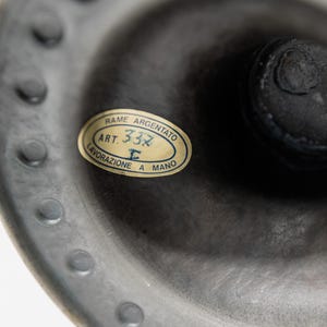 May include: Close-up of a dark gray metal object with a label. The label is oval-shaped with the text "RAME ARGENTATO ART. 332 LAVORAZIONE A MANO". The object has a textured surface and a series of small, evenly spaced bumps around the edge.