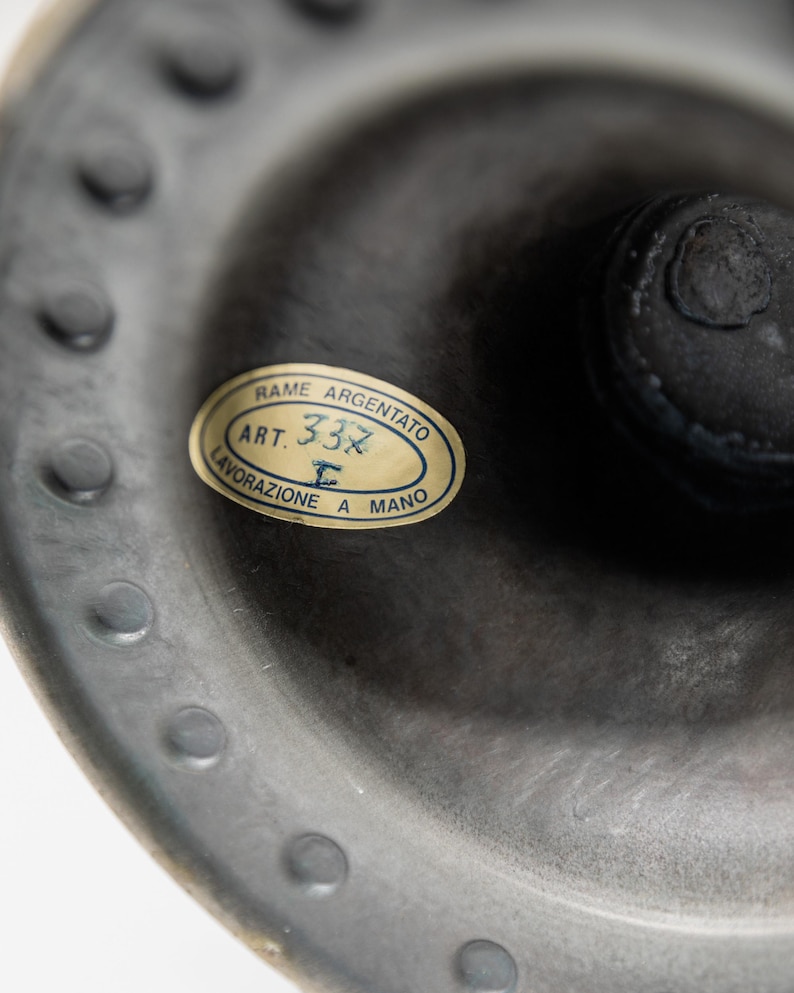 May include: Close-up of a dark gray metal object with a label. The label is oval-shaped with the text "RAME ARGENTATO ART. 332 LAVORAZIONE A MANO". The object has a textured surface and a series of small, evenly spaced bumps around the edge.