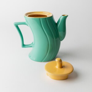 May include: A teal ceramic teapot with a curved body and a yellow interior. The teapot has a matching yellow lid and a unique handle design. The teapot is a decorative kitchen item.