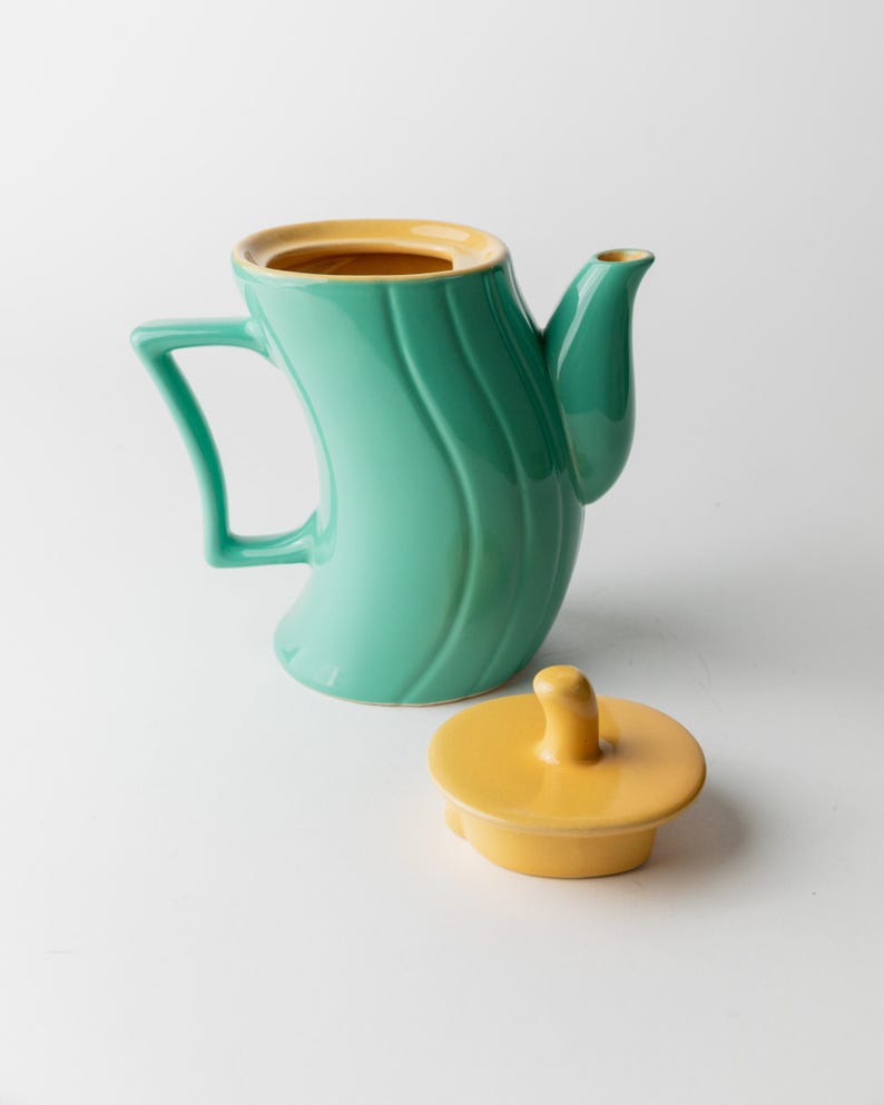 May include: A teal ceramic teapot with a curved body and a yellow interior. The teapot has a matching yellow lid and a unique handle design. The teapot is a decorative kitchen item.