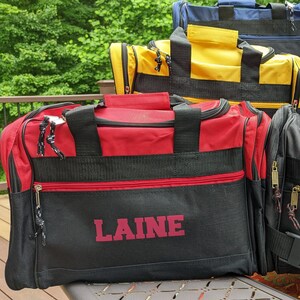 K&ouml;nnte beinhalten: Eine schwarze und rote Reisetasche mit dem Namen "LAINE" in rot auf der Vorderseite. Die Tasche hat schwarze Tr&auml;ger und einen Rei&szlig;verschluss.