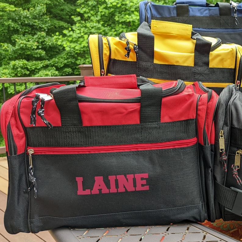 K&ouml;nnte beinhalten: Eine schwarze und rote Reisetasche mit dem Namen "LAINE" in rot auf der Vorderseite. Die Tasche hat schwarze Tr&auml;ger und einen Rei&szlig;verschluss.