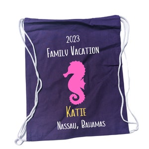 May include: A navy blue drawstring backpack with white drawstrings. The bag features the text "2023 Family Vacation" and a pink seahorse graphic. Below the seahorse is the name "Katie" in yellow, and "Nassau, Bahamas" in white.