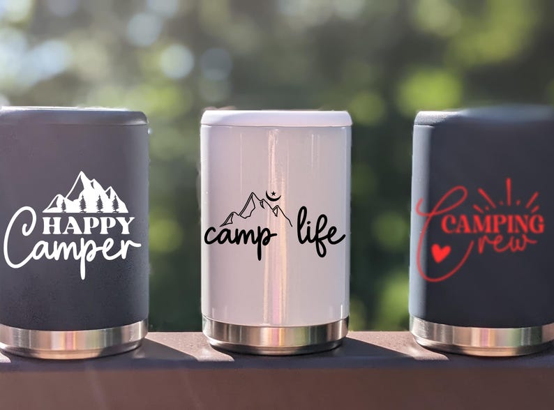 Puede incluir: Tres contenedores de bebidas aislados con diferentes dise&ntilde;os. El primer contenedor es negro con texto blanco que dice "HAPPY Camper" y un gr&aacute;fico de cordillera. El segundo contenedor es blanco con texto negro que dice "camp life" y un gr&aacute;fico de cordillera. El tercer contenedor es negro con texto rojo que dice "CAMPING crew" y un gr&aacute;fico de coraz&oacute;n.