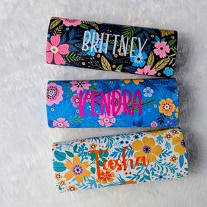 Peut inclure: Trois étuis à imprimé floral avec des noms personnalisés. L'étui supérieur est noir avec des fleurs roses et bleues et le nom "Brittney" en blanc. L'étui du milieu est bleu avec des fleurs roses et le nom "Kendra" en rose. L'étui inférieur est blanc avec des fleurs jaunes et oranges et le nom "Tasha" en orange.