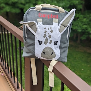 May include: A gray backpack with a white giraffe face on the front. The backpack has a zippered pocket and a name tag that says "Campbell".