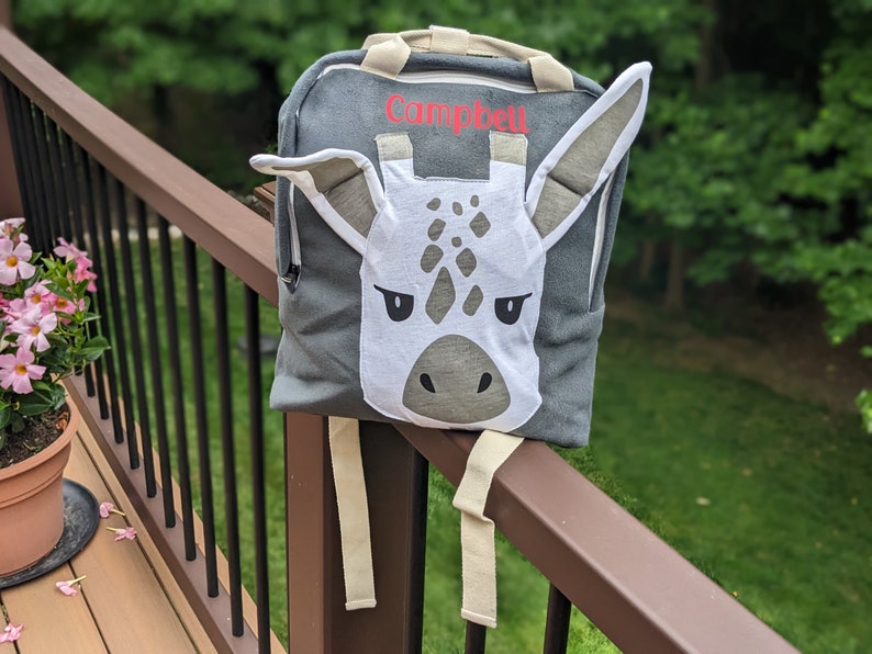 May include: A gray backpack with a white giraffe face on the front. The backpack has a zippered pocket and a name tag that says "Campbell".