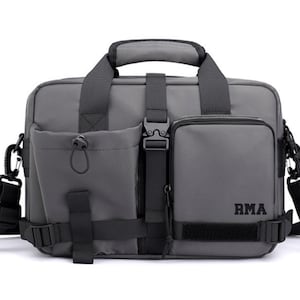 May include: A gray messenger bag with black straps and buckles. The bag has a large front pocket with a zipper and a smaller pocket with the text "RMA" on it.